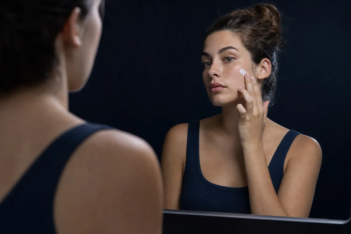 A young woman in a navy tank top dabbing sunscreen onto her cheekbone in a mirror