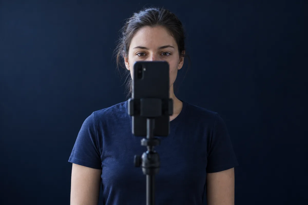 Young woman taking a front-facing progress photo with a phone on a tripod by a window