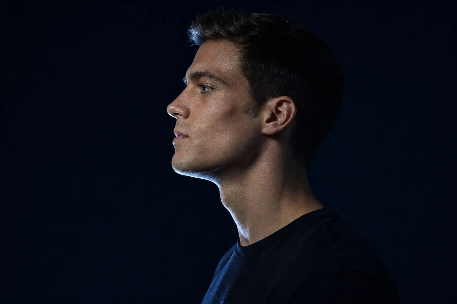 Side profile of a man with a defined jawline, lit from behind against a dark background